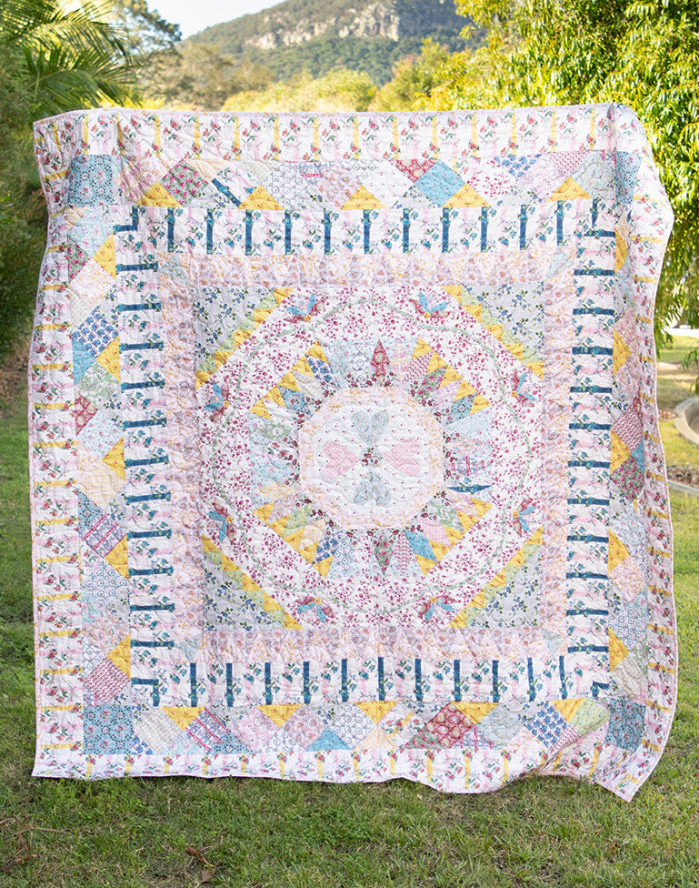 A large patchwork quilt with a detailed mandala pattern in pastel shades of pink, blue, yellow, and white, hanging outdoors on a grassy lawn with trees and a mountain in the background.