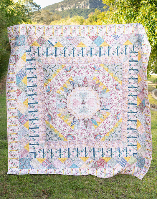 A large patchwork quilt with a detailed mandala pattern in pastel shades of pink, blue, yellow, and white, hanging outdoors on a grassy lawn with trees and a mountain in the background.